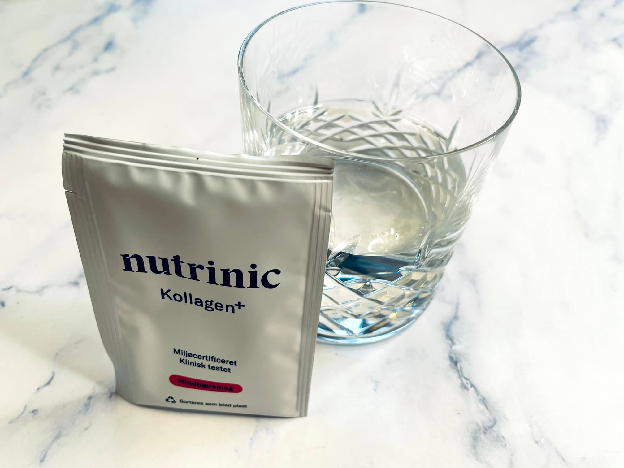 Doseringsbrev med Nutrinic Kollagen+ med hindbærsmag foran glas med vand